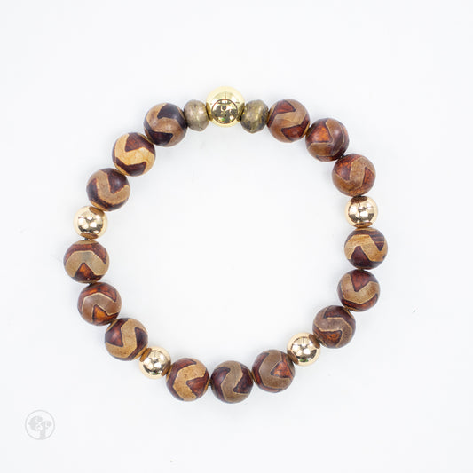 Top view of a brown chevron bead bracelet with tan markings, reflective gold-tone round accents, and textured metallic spacers beside an emblem bead.